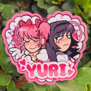 May include: A pink heart-shaped sticker featuring two anime-style characters. One character has pink hair and the other has purple hair. The word "YURI" is written in pink letters with star accents.