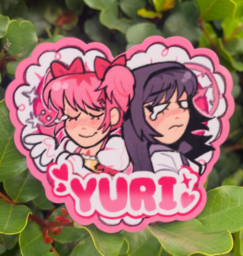 May include: A pink heart-shaped sticker featuring two anime-style characters. One character has pink hair and the other has purple hair. The word "YURI" is written in pink letters with star accents.