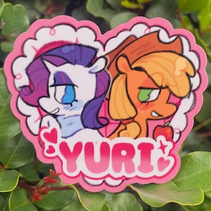 May include: A heart-shaped sticker featuring two cartoon ponies. One pony is white with purple hair, the other is orange with a green eye. The word "YURI" is written in pink letters at the bottom of the sticker. The sticker has a pink scalloped border.