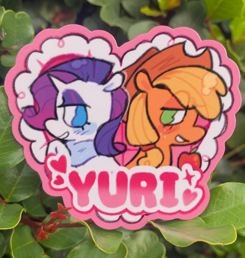 May include: A heart-shaped sticker featuring two cartoon ponies. One pony is white with purple hair, the other is orange with a green eye. The word "YURI" is written in pink letters at the bottom of the sticker. The sticker has a pink scalloped border.