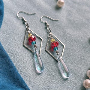 May include: A pair of silver-toned dangle earrings featuring a diamond-shaped metal frame. Each earring is accented with a small yellow bead, a faceted red bead, a small blue bead, and a clear, iridescent, teardrop-shaped glass pendant.