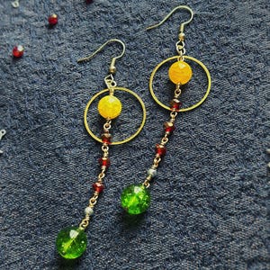 May include: A pair of dangle earrings featuring gold-tone metal hooks and thin chains. Each earring includes a circular gold-tone frame, a faceted yellow bead, several small dark red beads, and a large, round, faceted green glass bead at the base.