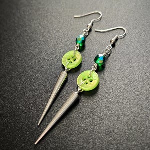 May include: A pair of handmade earrings with a unique design. Each earring features a silver-tone spike pendant, a bright green button, and green faceted beads. The earrings hang from silver-tone ear wires, creating a striking contrast against the dark background.