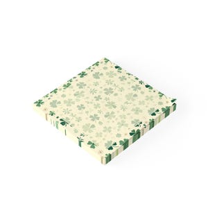 May include: Square notepad with a cream-colored background and a pattern of green clovers and floral elements. The edges are decorated with green stripes and clover designs, perfect for St. Patrick's Day or nature-themed notes.
