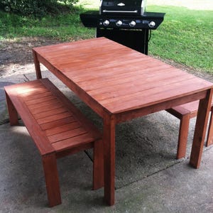 May include: A wooden outdoor dining set with a rectangular table and two benches. The table and benches are made of stained wood and have a natural wood grain. The set is on a concrete patio.