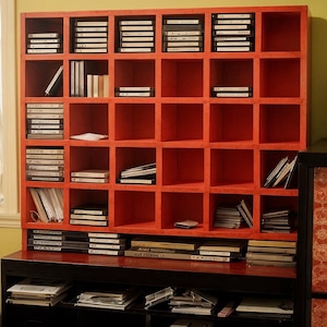 May include: A red wooden storage unit with multiple square compartments, filled with various media including CDs and cassette tapes. The unit sits atop a black cabinet, creating a functional and organized storage solution for media collections.