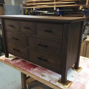 Op de afbeelding: Een donkerbruine houten dressoir met negen laden en zwarte metalen ladegrepen. De dressoir staat op een houten oppervlak.