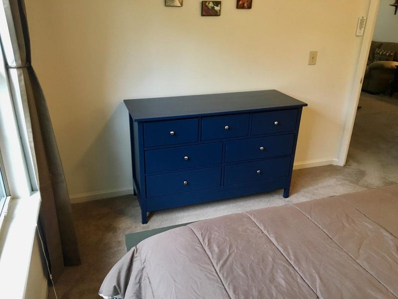 May include: A dark blue dresser with six drawers and silver drawer pulls. The dresser is in a bedroom setting with a tan comforter on the bed.