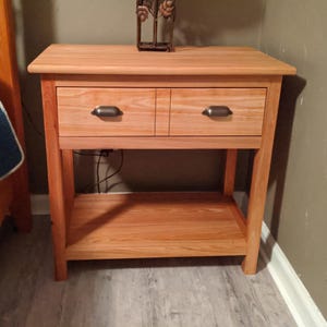 Pode incluir: Uma mesa de cabeceira de madeira com duas gavetas e uma prateleira inferior. As gavetas têm puxadores de metal. A mesa de cabeceira é feita de madeira clara com um acabamento natural.