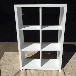May include: White cube shelf unit with six open compartments. The shelf is made of wood and has a simple, modern design.