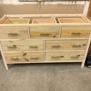Op de afbeelding: Een houten dressoir met zes laden en gouden handvatten. Het dressoir is gemaakt van onbehandeld hout en heeft een eenvoudig ontwerp.