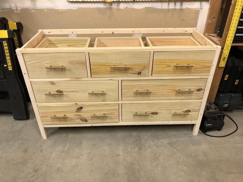Op de afbeelding: Een houten dressoir met zes laden en gouden handvatten. Het dressoir is gemaakt van onbehandeld hout en heeft een eenvoudig ontwerp.