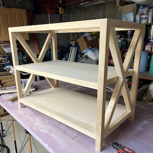 Puede incluir: Estantería de madera sin terminar con dos estantes y un diseño en forma de X en los laterales. La estantería está hecha de madera de color claro y está sobre una superficie morada. Se ve un taladro y otras herramientas.