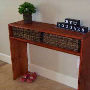 Może przedstawiać: Drewniany stolik konsolowy z ciemnobrązowym wykończeniem. Stolik ma dwa wiklinowe kosze do przechowywania i tabliczkę z napisem "BYU COUGARS".