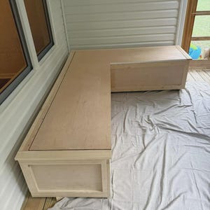 May include: L-shaped wooden storage bench, unfinished, with a light wood grain. The bench is constructed from rectangular panels and features a simple, modern design. It is placed on a wooden deck covered with a gray sheet.