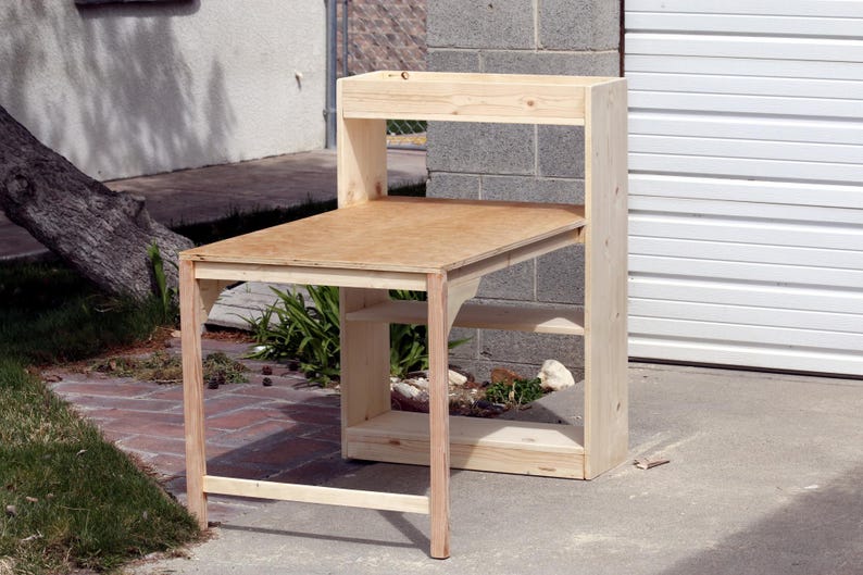 May include: A wooden desk with a light brown tabletop and a shelf. The desk has a natural wood finish and is supported by four legs. The desk also has a shelf for storage. The desk is approximately 40 inches wide and 30 inches deep.