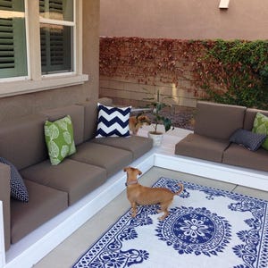 May include: A white wooden patio sectional sofa with brown cushions and blue and green throw pillows. A small brown dog is standing on a white and blue patterned rug in front of the sofa.