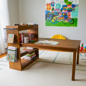 May include: A wooden desk with attached shelving and storage baskets. The desk has a dark brown finish and is set on a patterned rug. Books, art supplies, and an open book are visible on the desk. A colorful painting hangs on the wall.