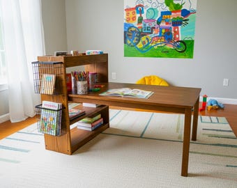 Op de afbeelding: Een houten bureau met ingebouwde planken en draadmandjes. Het bureau heeft een donkerbruine afwerking en staat op een patroonkleed. Boeken, kunstbenodigdheden en een open boek liggen op het bureau. Een kleurrijk schilderij hangt aan de muur.