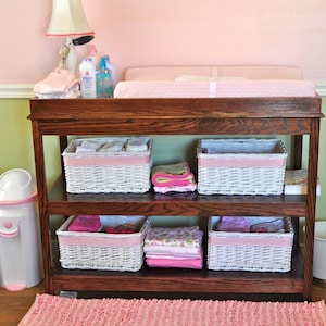 May include: A wooden changing table with four white wicker baskets. The baskets are filled with baby clothes and other items. The changing table is in a nursery with pink walls and a green accent wall.