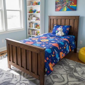 May include: A wooden twin-size bed with a dark brown finish. The bed is made up with a blue comforter and pillow set featuring a colorful space-themed design. A white teddy bear sits on the pillow. The room has a bookshelf and a gray rug.