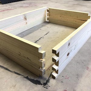 Op de afbeelding: Vier stukken onbehandeld hout met in elkaar grijpende verbindingen, klaar om in elkaar gezet te worden tot een doos. Het hout is lichtbruin en heeft een glad oppervlak. De verbindingen zijn genummerd.