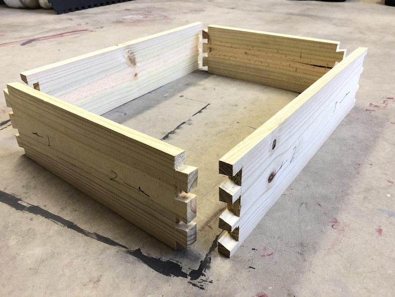 Op de afbeelding: Vier stukken onbehandeld hout met in elkaar grijpende verbindingen, klaar om in elkaar gezet te worden tot een doos. Het hout is lichtbruin en heeft een glad oppervlak. De verbindingen zijn genummerd.