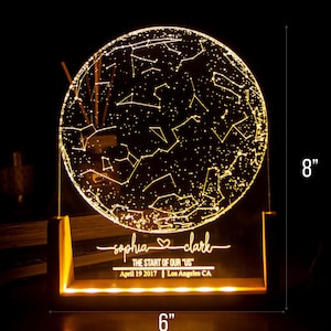 Puede incluir: Una luz nocturna personalizada de mapa de estrellas con una base de acr&iacute;lico transparente y un marco de madera. La luz muestra la constelaci&oacute;n del cielo nocturno del 19 de abril de 2017 en Los &Aacute;ngeles, California. Los nombres "sophia" y "clark" est&aacute;n grabados en la base, junto con el texto "The start of our "US"". La luz mide 20,3 cm de alto y 15,2 cm de ancho.