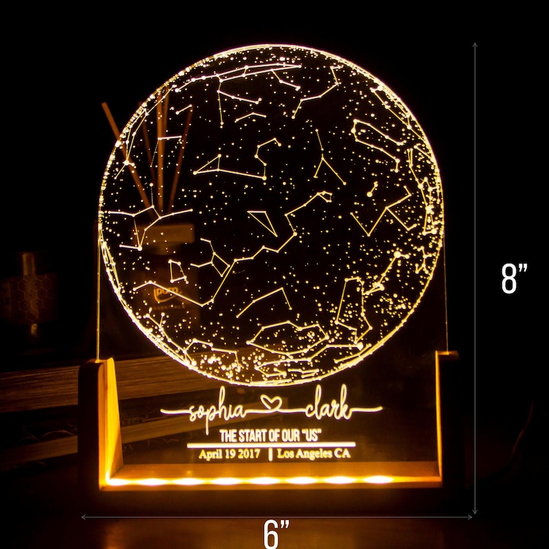 Puede incluir: Una luz nocturna personalizada de mapa de estrellas con una base de acr&iacute;lico transparente y un marco de madera. La luz muestra la constelaci&oacute;n del cielo nocturno del 19 de abril de 2017 en Los &Aacute;ngeles, California. Los nombres "sophia" y "clark" est&aacute;n grabados en la base, junto con el texto "The start of our "US"". La luz mide 20,3 cm de alto y 15,2 cm de ancho.