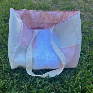 May include: A pink and white patchwork tote bag with a white lace trim and a pink satin lining. The bag has a white label that says "Handmade by Me".