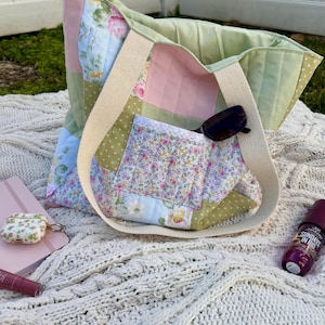 May include: A patchwork tote bag with a cream-colored strap, featuring floral and pastel fabric squares. The bag contains sunglasses and a floral pocket. Other items include a pink notebook, a floral case, and a bottle of lotion, all on a cream-colored knitted blanket.