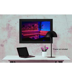 May include: A black framed print of a neon-lit cityscape with palm trees and a bar scene. A black desk lamp, a laptop, and a small white vase with pink flowers are on a white desk. The text "Frame not included" is visible.