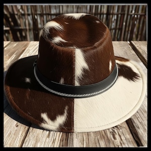 May include: A two-tone cowboy hat with a brown and white cowhide pattern. The hat features a black leather band with a silver-colored trim. The brim is wide and the crown is rounded, typical of a western-style hat.