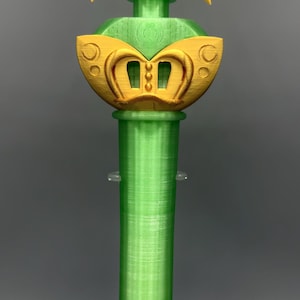 K&ouml;nnte beinhalten: Ein gr&uuml;ner und gelber Kunststoffstab mit einer sternf&ouml;rmigen Spitze und einem zentralen kreisf&ouml;rmigen Emblem, das die Zahl 21 zeigt. Der Stab hat einen zylindrischen Griff und einen dekorativen goldfarbenen Abschnitt unter dem Stern, alles mit sichtbarer 3D-Druckstruktur.