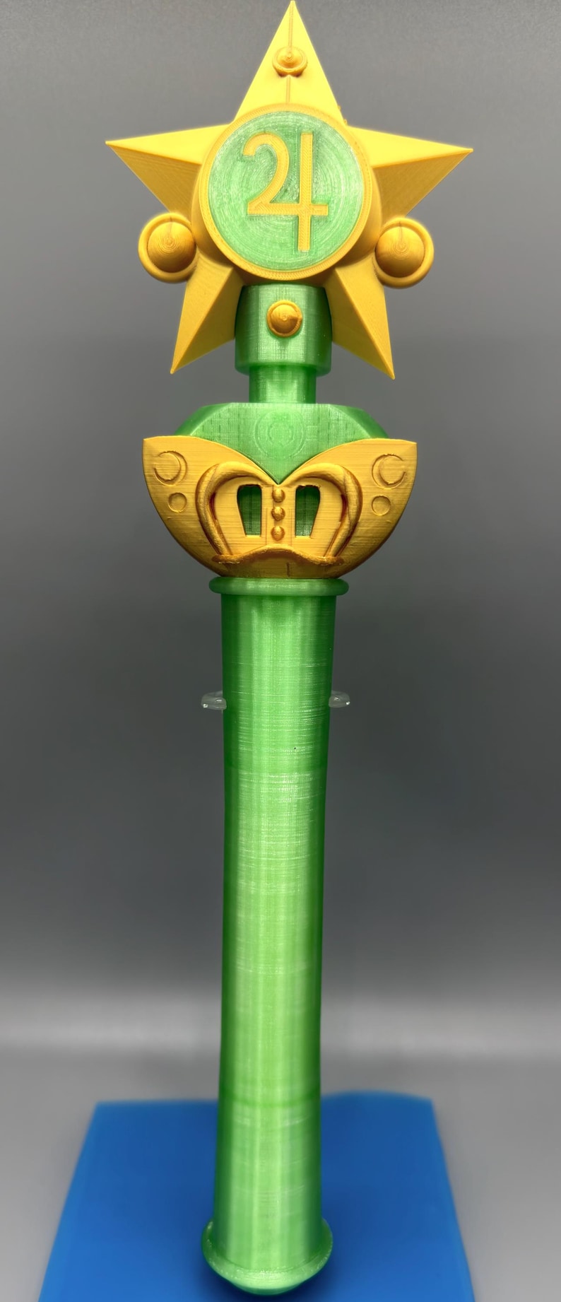 K&ouml;nnte beinhalten: Ein gr&uuml;ner und gelber Kunststoffstab mit einer sternf&ouml;rmigen Spitze und einem zentralen kreisf&ouml;rmigen Emblem, das die Zahl 21 zeigt. Der Stab hat einen zylindrischen Griff und einen dekorativen goldfarbenen Abschnitt unter dem Stern, alles mit sichtbarer 3D-Druckstruktur.