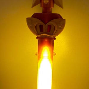 K&ouml;nnte beinhalten: Ein vertikaler, stabartiger Gegenstand mit einem leuchtenden, durchscheinenden orangefarbenen zylindrischen Schaft. Der obere Teil ist mit einem goldfarbenen Sternemblem verziert, das den Buchstaben P enth&auml;lt und &uuml;ber einem fl&uuml;gelf&ouml;rmigen Element sitzt.
