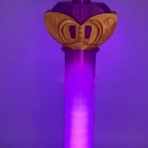 K&ouml;nnte beinhalten: Ein vertikaler Zauberstab mit einem leuchtend violetten zylindrischen Schaft, der an einem goldenen sternf&ouml;rmigen Aufsatz befestigt ist. Der Aufsatz enth&auml;lt ein zentrales violettes kreisf&ouml;rmiges Emblem mit einem goldenen Symbol und goldenen Akzenten. Der Stab steht auf einem quadratischen Sockel.