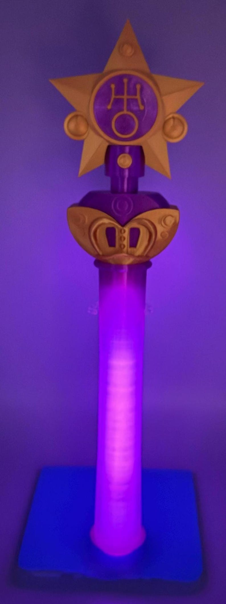 K&ouml;nnte beinhalten: Ein vertikaler Zauberstab mit einem leuchtend violetten zylindrischen Schaft, der an einem goldenen sternf&ouml;rmigen Aufsatz befestigt ist. Der Aufsatz enth&auml;lt ein zentrales violettes kreisf&ouml;rmiges Emblem mit einem goldenen Symbol und goldenen Akzenten. Der Stab steht auf einem quadratischen Sockel.