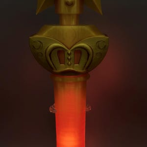 K&ouml;nnte beinhalten: Ein goldfarbener Requisitenstab mit einem gro&szlig;en Stern an der Spitze, der ein zentrales kreisf&ouml;rmiges Emblem mit einem Herz- und Kreuzsymbol zeigt. Unter dem Stern befindet sich ein dekorativer Sockel mit geschwungenen Details, der Griff leuchtet orange.