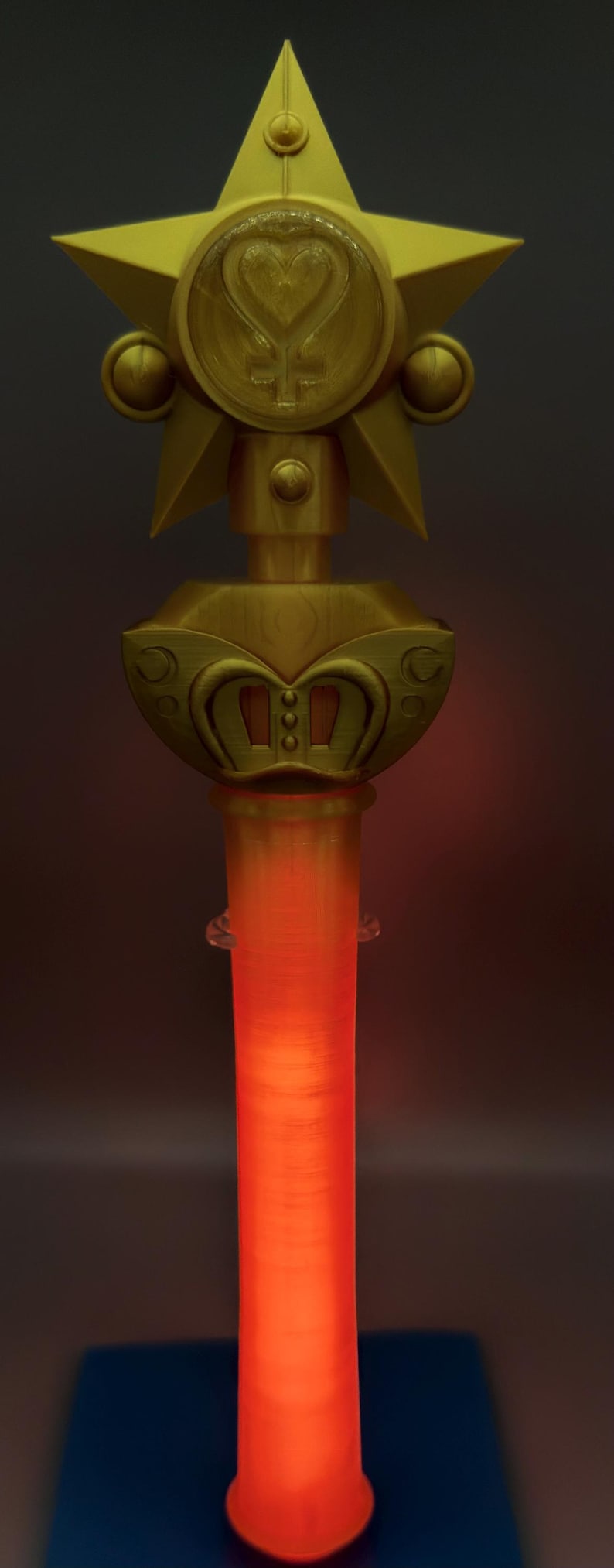 K&ouml;nnte beinhalten: Ein goldfarbener Requisitenstab mit einem gro&szlig;en Stern an der Spitze, der ein zentrales kreisf&ouml;rmiges Emblem mit einem Herz- und Kreuzsymbol zeigt. Unter dem Stern befindet sich ein dekorativer Sockel mit geschwungenen Details, der Griff leuchtet orange.