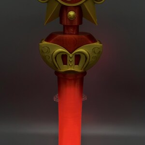 K&ouml;nnte beinhalten: Ein Zauberstab-Requisit mit einem goldenen Stern an der Spitze und einem roten, kreisf&ouml;rmigen Zentrum, das ein goldenes Mars-Symbol zeigt. Unter dem Stern befindet sich ein goldenes, kronenartiges Element mit roten Akzenten. Der Griff ist ein leuchtender, roter Zylinder.