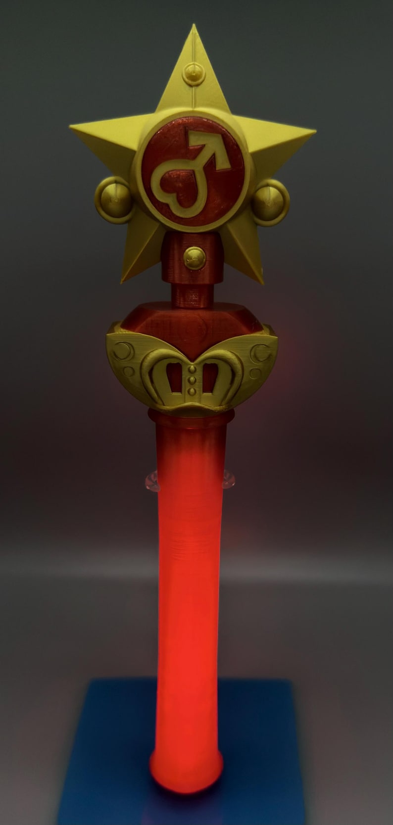 K&ouml;nnte beinhalten: Ein Zauberstab-Requisit mit einem goldenen Stern an der Spitze und einem roten, kreisf&ouml;rmigen Zentrum, das ein goldenes Mars-Symbol zeigt. Unter dem Stern befindet sich ein goldenes, kronenartiges Element mit roten Akzenten. Der Griff ist ein leuchtender, roter Zylinder.