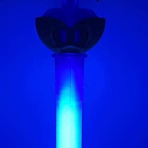 K&ouml;nnte beinhalten: Ein vertikaler Requisitenstab mit einem leuchtend hellblauen zylindrischen Griff und einem dunklen sternf&ouml;rmigen Aufsatz. In der Mitte des Sterns befindet sich ein kreisf&ouml;rmiges Emblem mit dem Saturnsymbol, einem Kreuz auf einer gebogenen Linie.