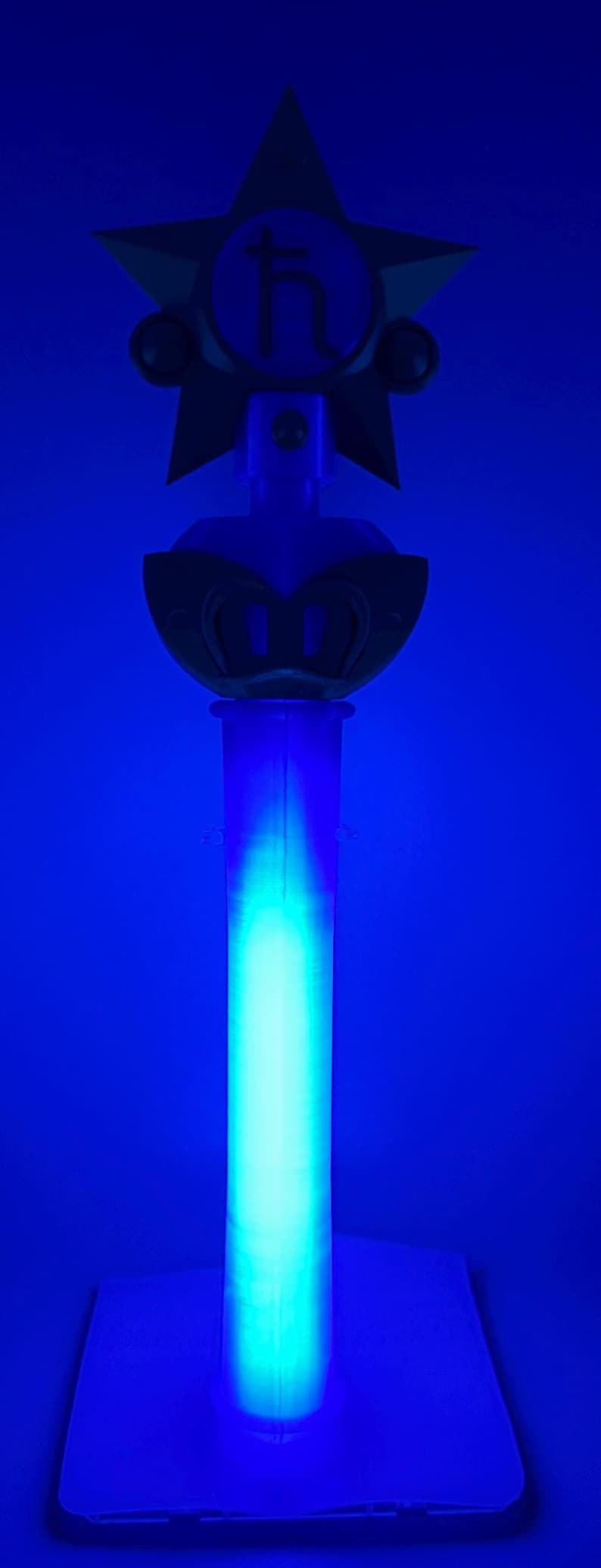 K&ouml;nnte beinhalten: Ein vertikaler Requisitenstab mit einem leuchtend hellblauen zylindrischen Griff und einem dunklen sternf&ouml;rmigen Aufsatz. In der Mitte des Sterns befindet sich ein kreisf&ouml;rmiges Emblem mit dem Saturnsymbol, einem Kreuz auf einer gebogenen Linie.
