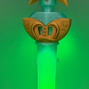 K&ouml;nnte beinhalten: Ein Requisitenstab mit einem goldenen Stern an der Spitze und einem hellblauen kreisf&ouml;rmigen Emblem mit einem Dreizacksymbol. Unter dem Stern befindet sich eine hellblaue Kugel mit einem goldenen Dekorelement. Der Griff ist ein durchscheinender hellgr&uuml;ner Zylinder.