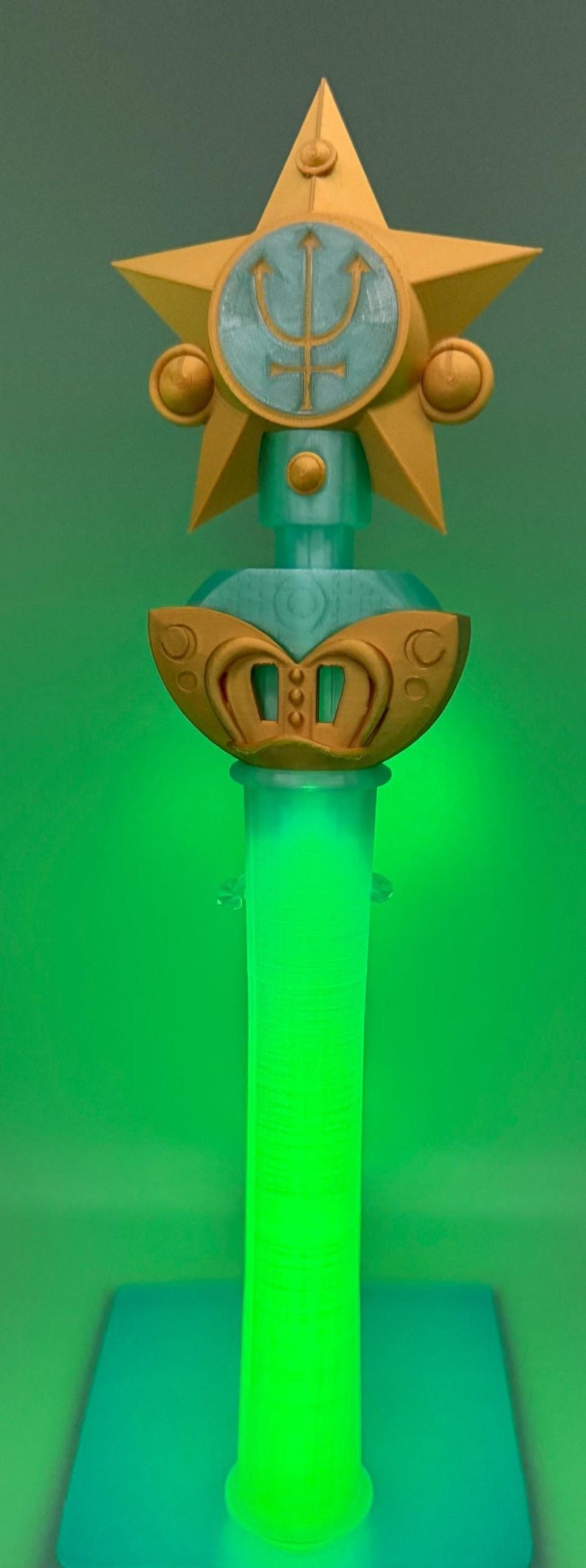 K&ouml;nnte beinhalten: Ein Requisitenstab mit einem goldenen Stern an der Spitze und einem hellblauen kreisf&ouml;rmigen Emblem mit einem Dreizacksymbol. Unter dem Stern befindet sich eine hellblaue Kugel mit einem goldenen Dekorelement. Der Griff ist ein durchscheinender hellgr&uuml;ner Zylinder.