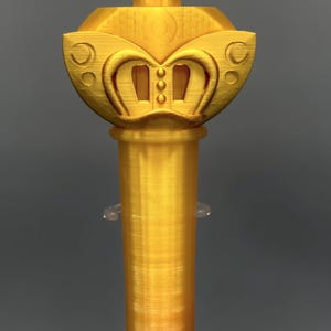 K&ouml;nnte beinhalten: Ein goldfarbener, 3D-gedruckter Zauberstab mit einem gro&szlig;en f&uuml;nfzackigen Stern an der Spitze, der ein zentrales kreisf&ouml;rmiges Emblem mit einem Herz- und Kreuzsymbol aufweist. Der Griff ist zylindrisch und glatt, mit geschwungenen Details.