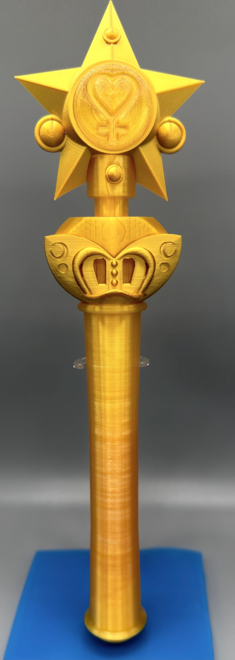 K&ouml;nnte beinhalten: Ein goldfarbener, 3D-gedruckter Zauberstab mit einem gro&szlig;en f&uuml;nfzackigen Stern an der Spitze, der ein zentrales kreisf&ouml;rmiges Emblem mit einem Herz- und Kreuzsymbol aufweist. Der Griff ist zylindrisch und glatt, mit geschwungenen Details.