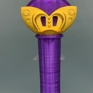 K&ouml;nnte beinhalten: Ein 3D-gedruckter Requisitenstab mit einem gelben Stern an der Spitze und einem violetten kreisf&ouml;rmigen Emblem in der Mitte. Der Stab hat einen violetten zylindrischen Griff und einen gelben dekorativen Basisabschnitt mit drei vertikalen Schlitzen.