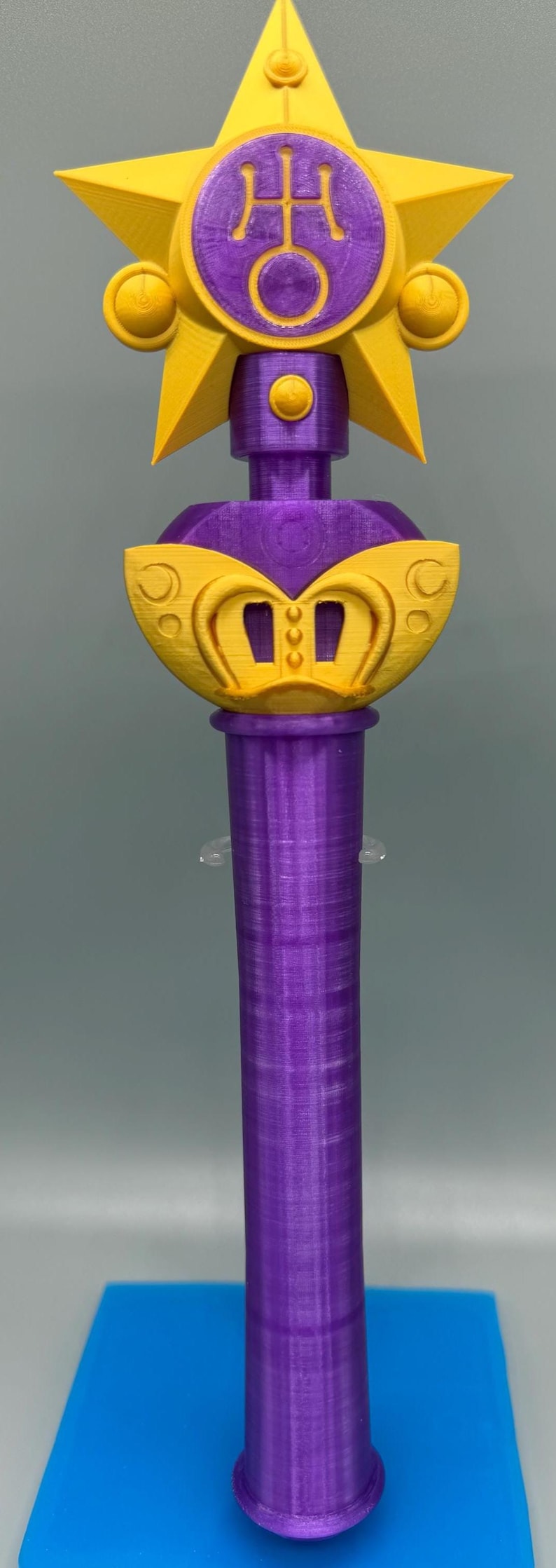 K&ouml;nnte beinhalten: Ein 3D-gedruckter Requisitenstab mit einem gelben Stern an der Spitze und einem violetten kreisf&ouml;rmigen Emblem in der Mitte. Der Stab hat einen violetten zylindrischen Griff und einen gelben dekorativen Basisabschnitt mit drei vertikalen Schlitzen.